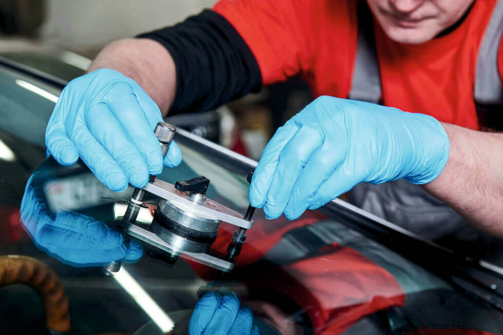 Una foto ravvicinata di una piccola scheggiatura a forma di stella sul parabrezza di un'auto, vista dall'interno. Sullo sfondo sfocato si intravede una strada con altre auto in transito, a simboleggiare l'importanza di una riparazione tempestiva per la sicurezza stradale. La scheggiatura è al centro dell'attenzione, con il resto dell'immagine che suggerisce il contesto di un viaggio interrotto.